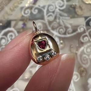 Könnte beinhalten: Goldfarbener Anhänger mit einem herzförmigen Rubin-Edelstein in der Mitte. Der Anhänger hat ein Retro-Design mit drei kleinen, runden, klaren Edelsteinen unterhalb des Rubins. Der Anhänger wird zwischen zwei Fingern gehalten.