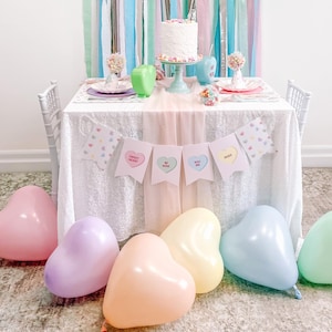 Peut inclure: Une table de fête de la Saint-Valentin avec une palette de couleurs pastel. La table est décorée d'une bannière qui dit "Sweet Heart, Be Mine, Kiss Me, Hugs". Il y a des ballons en forme de cœur dans des couleurs pastel sur le sol.