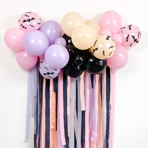 Peut inclure: Une d&eacute;coration de f&ecirc;te d'Halloween avec une guirlande de ballons violets, roses et noirs avec des silhouettes de chauves-souris. Les ballons sont suspendus &agrave; un mur avec un rideau de rubans roses, violets et noirs.