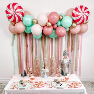 Peut inclure: Une table de Noël festive avec une nappe blanche, ornée d'accents roses, vert menthe et dorés. La décoration comprend une arche de ballons, des ballons cannes de bonbon, des casse-noisettes et des coussins moelleux. Des assiettes et des gobelets sont dressés pour un repas de fête.