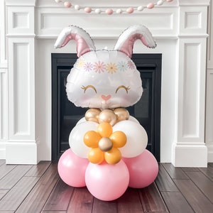 May include: An Easter-themed balloon display. A large bunny head balloon with a floral crown sits above a base of pink, white, and gold balloons. An orange flower balloon is also included. Ideal for Easter celebrations.