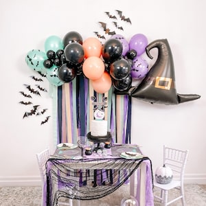 May include: A Halloween-themed table setting with a black and white spiderweb tablecloth, a white cake with a black spider on top, and a black witch hat balloon. The table is decorated with black, purple, and orange balloons, and there are black bats hanging from the wall.