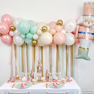 May include: A festive party backdrop featuring a pink, white and green balloon garland, gold ribbon streamers and a large pink and green Nutcracker balloon. A table is set with pink and white plates, gold cutlery and a Nutcracker-themed party decoration.