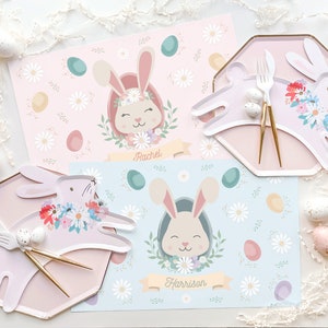 May include: Two pastel-colored placemats with bunny illustrations and floral accents. The placemats feature the names "Rachel" and "Harrison".
