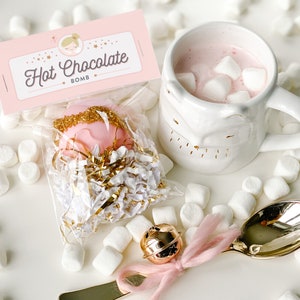 May include: A pink hot chocolate bomb in a clear bag with gold sprinkles, next to a white mug filled with pink hot chocolate and marshmallows. The mug has a face design. A gold spoon with a pink ribbon and a small gold bell are also present.
