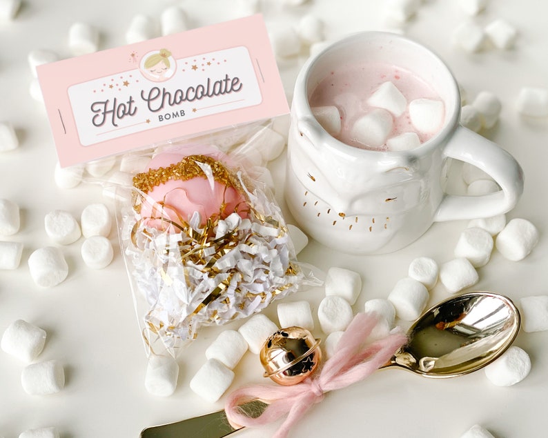 May include: A pink hot chocolate bomb in a clear bag with gold sprinkles, next to a white mug filled with pink hot chocolate and marshmallows. The mug has a face design. A gold spoon with a pink ribbon and a small gold bell are also present.