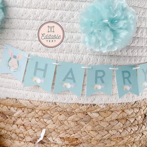 May include: A light blue banner with the name "HARRY" spelled out in white letters. The banner is decorated with pastel Easter eggs and a bunny rabbit illustration. The banner is hanging from a white wicker basket with a light blue pom-pom on top.