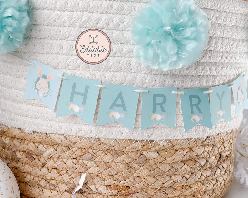 May include: A light blue banner with the name "HARRY" spelled out in white letters. The banner is decorated with pastel Easter eggs and a bunny rabbit illustration. The banner is hanging from a white wicker basket with a light blue pom-pom on top.