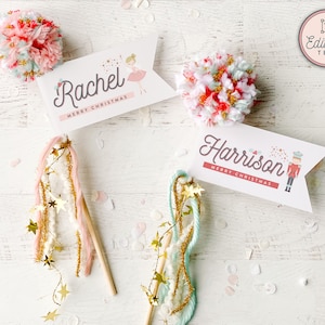 Puede incluir: Dos etiquetas de regalo de Navidad con un pompón rosa y blanco en la parte superior de cada una. La etiqueta de la izquierda tiene una ilustración de bailarina y dice "Rachel Feliz Navidad". La etiqueta de la derecha tiene una ilustración de cascanueces y dice "Harrison Feliz Navidad".