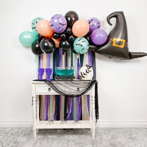 May include: A Halloween party table setting with a witch hat balloon, purple, pink, and green balloons, a bowl of blue punch, and a white pumpkin with bats.