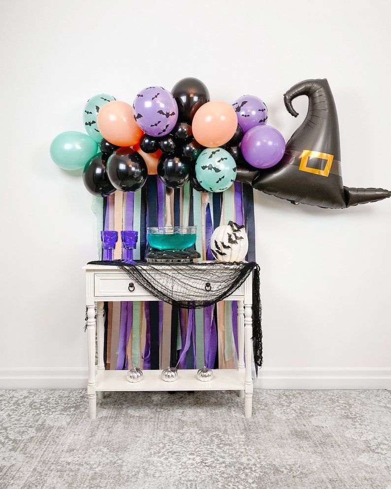 May include: A Halloween party table setting with a witch hat balloon, purple, pink, and green balloons, a bowl of blue punch, and a white pumpkin with bats.