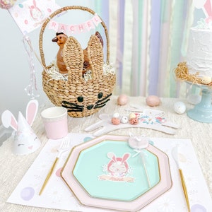 May include: A pastel-colored Easter table setting with a bunny-shaped wicker basket, a pink and green paper plate with a bunny illustration, and a bunny-shaped party hat. The table is decorated with a white tablecloth and a pink and white floral runner.
