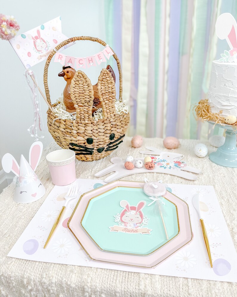 May include: A pastel-colored Easter table setting with a bunny-shaped wicker basket, a pink and green paper plate with a bunny illustration, and a bunny-shaped party hat. The table is decorated with a white tablecloth and a pink and white floral runner.