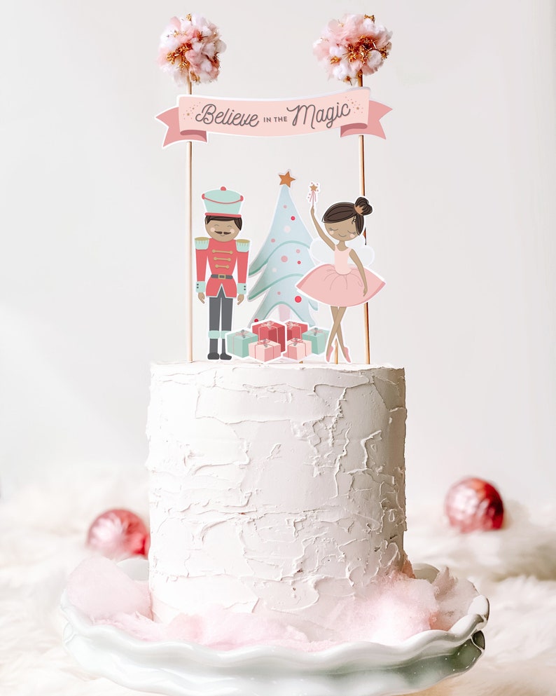 May include: A white cake decorated with a pink banner that says "Believe in the Magic". The cake is topped with a nutcracker, a ballerina, a Christmas tree, and presents. The cake is on a white cake stand with pink cotton candy around the base.
