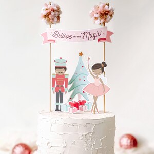 Pode incluir: Um bolo branco com uma &aacute;rvore de Natal rosa e verde, um quebra-nozes e uma bailarina no topo. O bolo &eacute; decorado com uma faixa que diz "Acredite na magia".