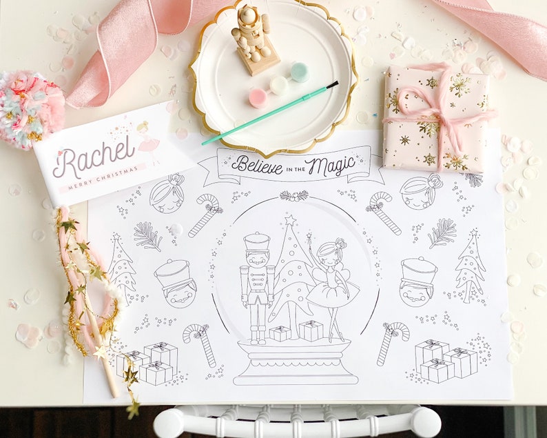 May include: A festive Christmas-themed scene with a coloring placemat featuring a snow globe design and the text "Believe in the Magic." Includes a place card, wrapped gift, paint palette, and a wooden nutcracker figurine. Pink and gold accents.