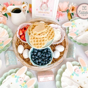 Pu&ograve; includere: Una tavola apparecchiata per un brunch con una tavolozza di colori pastello. La tavola &egrave; decorata con piatti, tovaglioli e un centrotavola a tema coniglietto, con waffle, frutta e mirtilli. Il centrotavola &egrave; circondato da fiori rosa e verdi pastello. La tavola include anche un cartello che dice "Testo modificabile".