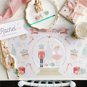 May include: A festive Christmas-themed table setting with a placemat featuring a snow globe design, a ballerina, and a nutcracker. Includes a personalized banner, wrapped gifts, and a paper cup with a straw.