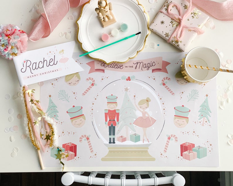May include: A festive Christmas-themed table setting with a placemat featuring a snow globe design, a ballerina, and a nutcracker. Includes a personalized banner, wrapped gifts, and a paper cup with a straw.
