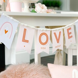 May include: A pink and white banner with the word "LOVE" in orange letters. The banner has a white fringe and is decorated with small hearts and rainbows. The banner says "LOVE YOU MOST" on the flags.
