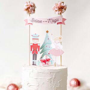 Puede incluir: Un pastel blanco con un adorno rosa y dorado que presenta un cascanueces, una bailarina, un &aacute;rbol de Navidad y regalos. El adorno tambi&eacute;n incluye una pancarta rosa con el texto "Believe in the Magic".
