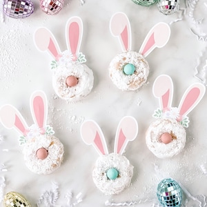 May include: Four white frosted donuts decorated with pink bunny ears and pastel flowers. Each donut has a pastel blue candy egg in the center.