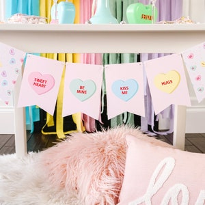 May include: A pink and white Valentine's Day banner with the words "Sweet Heart", "Be Mine", "Kiss Me", and "Hugs" printed on each flag. The banner is hanging from a white table with a pink and white fuzzy rug underneath.