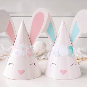 May include: Two Easter bunny cone hats. One hat features a floral design, the other has Easter eggs. Both hats have pink hearts, smiling faces, and large bunny ears with pink and blue accents. The hats are on a white surface.