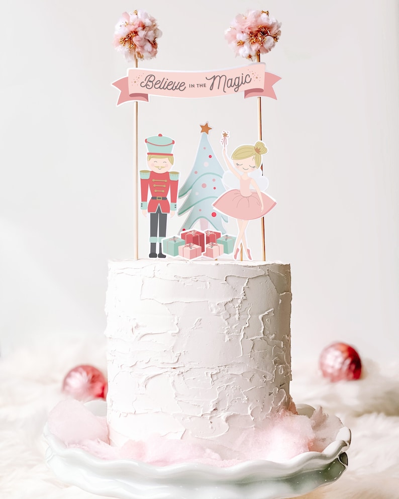 Puede incluir: Un pastel blanco con glaseado rosa y una decoraci&oacute;n navide&ntilde;a. La decoraci&oacute;n incluye un cascanueces, una bailarina, un &aacute;rbol de Navidad y regalos. La pancarta dice "Cree en la magia".