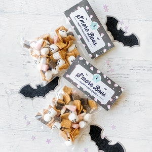 May include: Two clear plastic bags filled with a Halloween snack mix. The mix includes mini marshmallows, chocolate squares, graham cracker pieces, and candy corn. Each bag has a gray and white tag attached with the text "S'more Boos - Happy Halloween".