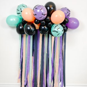 May include: A Halloween party backdrop with a balloon arch made of black, purple, teal, and orange balloons. The balloons are decorated with bats. The backdrop is made of streamers in various colors, including purple, teal, black, and peach.