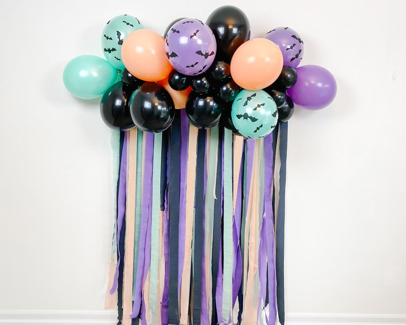 May include: A Halloween party backdrop with a balloon arch made of black, purple, teal, and orange balloons. The balloons are decorated with bats. The backdrop is made of streamers in various colors, including purple, teal, black, and peach.