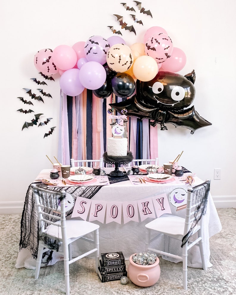 Peut inclure: Une table de f&ecirc;te d'Halloween avec une nappe rose, des chaises blanches et une banni&egrave;re qui dit "SPOOKY". La table est d&eacute;cor&eacute;e d'assiettes, de gobelets et de serviettes noirs et roses. Il y a aussi des ballons noirs et roses avec des chauves-souris imprim&eacute;es dessus. Un ballon de chauve-souris noir avec des yeux globuleux est accroch&eacute; au mur derri&egrave;re la table.
