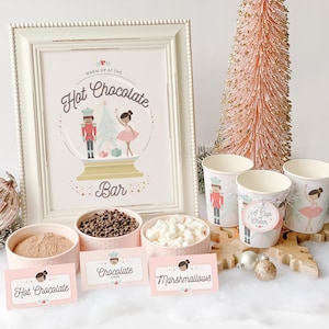 Puede incluir: Una barra de chocolate caliente rosa y blanca con un letrero que dice "Warm up at the Hot Chocolate Bar". El letrero presenta un globo de nieve con un cascanueces y una bailarina dentro. Hay tazones de mezcla de chocolate caliente, chispas de chocolate y malvaviscos. También hay vasos rosas y blancos con un diseño de cascanueces y bailarina.