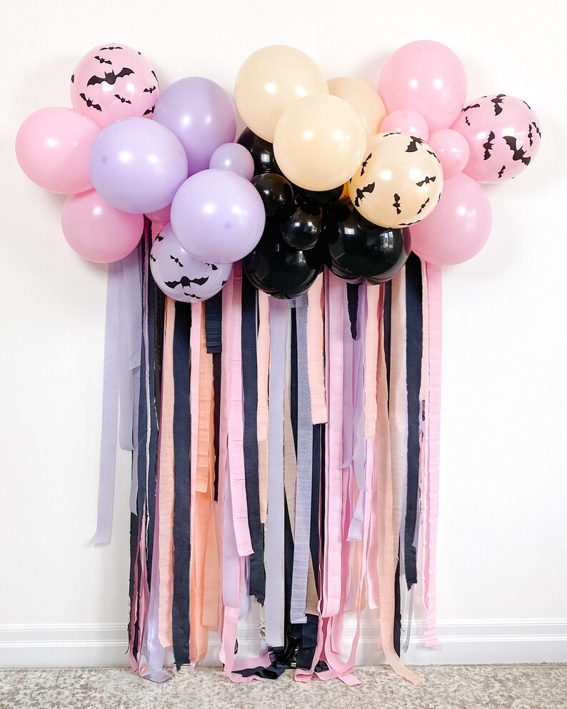 Peut inclure: Une d&eacute;coration de f&ecirc;te d'Halloween avec une guirlande de ballons violets, roses et noirs avec des silhouettes de chauves-souris. Les ballons sont suspendus &agrave; un mur avec un rideau de rubans roses, violets et noirs.