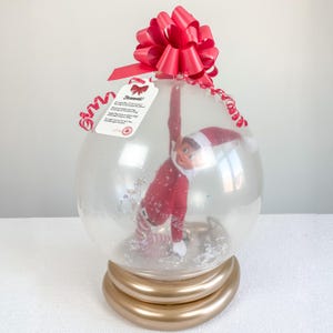 Peut inclure: Un globe à neige en plastique transparent avec une poupée d'elfe rouge et blanche à l'intérieur. Le globe est posé sur une base de ballon dorée et surmonté d'un nœud rouge. Le globe est muni d'une étiquette sur laquelle est inscrit "Farewell!" avec un message sur le départ de l'elfe pour le pôle Nord.