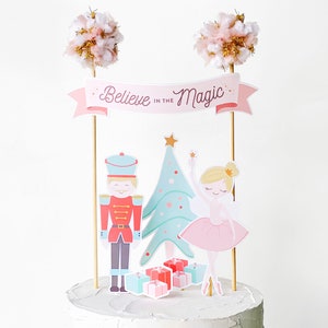 May include: A cake topper with a festive theme. It features a banner that reads "Believe in the Magic", a nutcracker, a Christmas tree, a ballerina, and presents. The topper is decorated with pink, gold, and light blue colors.