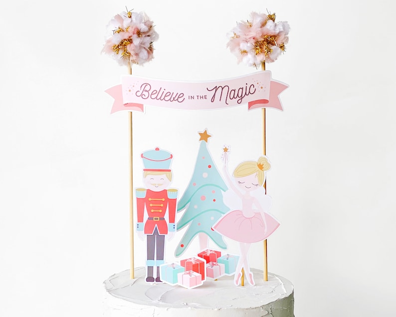 May include: A cake topper with a festive theme. It features a banner that reads "Believe in the Magic", a nutcracker, a Christmas tree, a ballerina, and presents. The topper is decorated with pink, gold, and light blue colors.