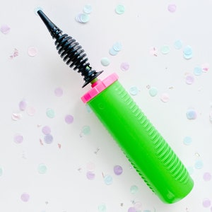 May include: A green balloon pump with a black nozzle and a pink handle. The pump is on a white surface with confetti.