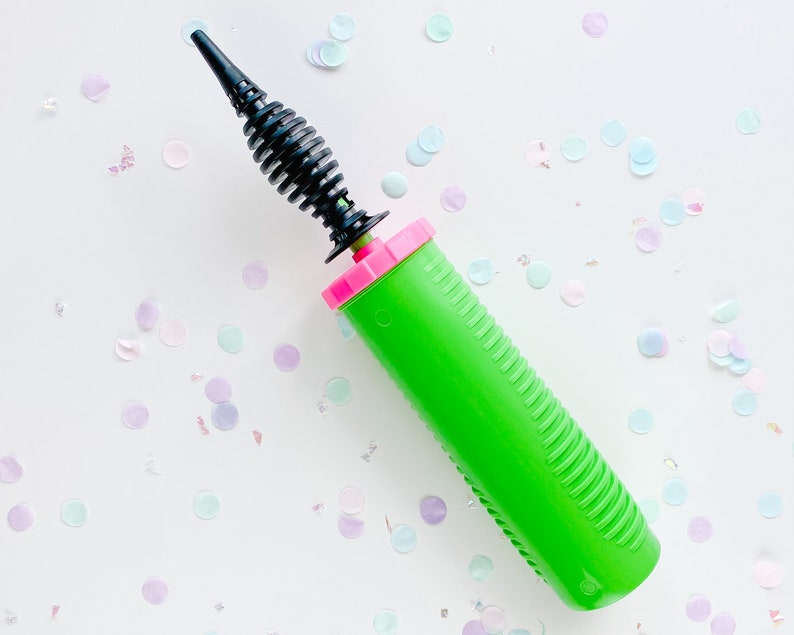 May include: A green balloon pump with a black nozzle and a pink handle. The pump is on a white surface with confetti.