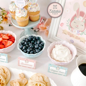 Pu&ograve; includere: Un cartello per un bar di waffle a tema coniglietto rosa e verde pastello con il testo "Waffle Bar" e una simpatica illustrazione di coniglietto. Il cartello &egrave; incorniciato di bianco ed &egrave; esposto su un tavolo con una variet&agrave; di condimenti per waffle, tra cui fragole, mirtilli, panna montata e caff&egrave;.