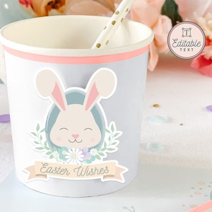 May include: A light blue paper cup with a pink rim features a sticker of a white bunny with a blue egg, green leaves, and flowers. The sticker also includes a banner with the text "Easter Wishes".