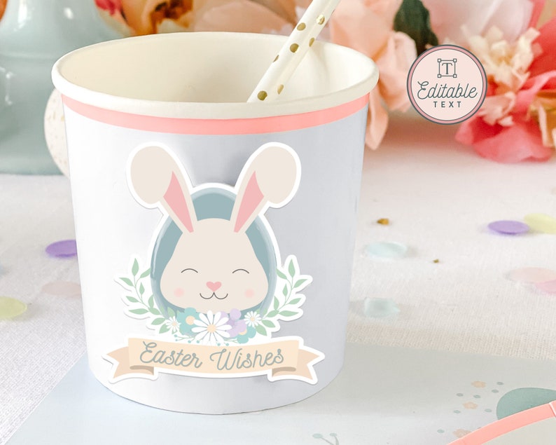 May include: A light blue paper cup with a pink rim features a sticker of a white bunny with a blue egg, green leaves, and flowers. The sticker also includes a banner with the text "Easter Wishes".