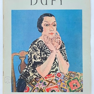 May include: A vintage book cover featuring the title "DUFY" in large, black capital letters. The cover displays a colorful painting of a woman with her hands clasped, wearing a patterned dress. The background is a solid blue.