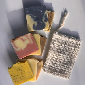 May include: A set of six handmade soap bars in various colors and patterns, including pink, yellow, and blue. The soaps are stacked on top of each other and are accompanied by a small white mesh bag.