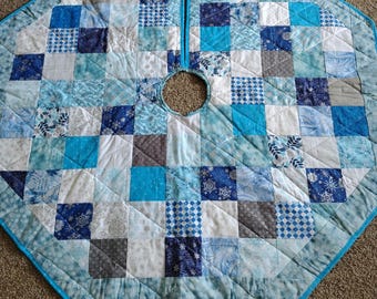 May include: A quilted Christmas tree skirt in shades of blue, white, and gray. The skirt is octagonal with a center opening and a split down the front. The patchwork design features various patterns, including snowflakes and geometric shapes.