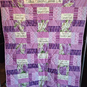 May include: A handmade quilt in shades of purple and white, featuring floral patterns and inspirational text. The quilt is displayed on a metal rack. The text includes phrases like "What Cancer Cannot Do" and other motivational statements.