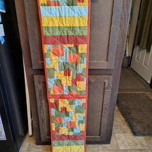 May include: A long, rectangular quilted table runner featuring a patchwork design of orange, yellow, green, and light blue fabrics. The runner includes sections of solid colours, polka dots, and floral patterns, all bordered by a solid orange fabric trim.
