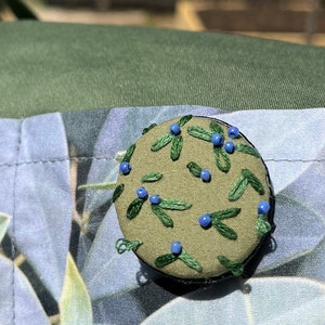 May include: A green fabric button with blue beads embroidered in a floral pattern.
