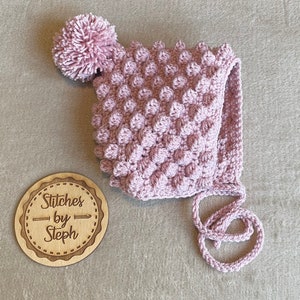 May include: A pink crocheted baby bonnet with a pom-pom on top and a tie at the bottom. The bonnet has a textured, bumpy design. The label "Stitches by Steph" is visible in the image.
